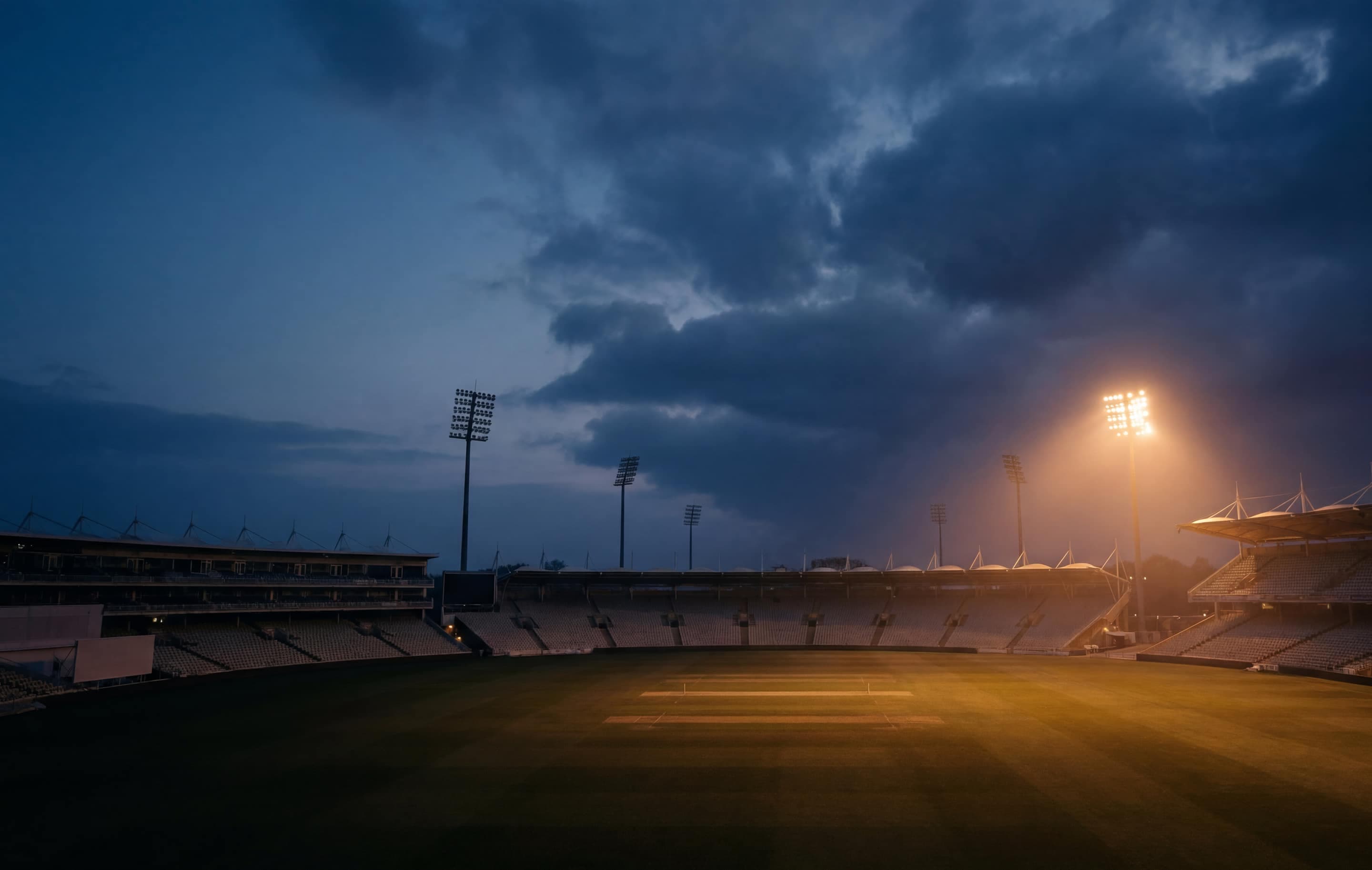 Floodlit cricket stadium at dusk, empty pitch under warm floodlights
